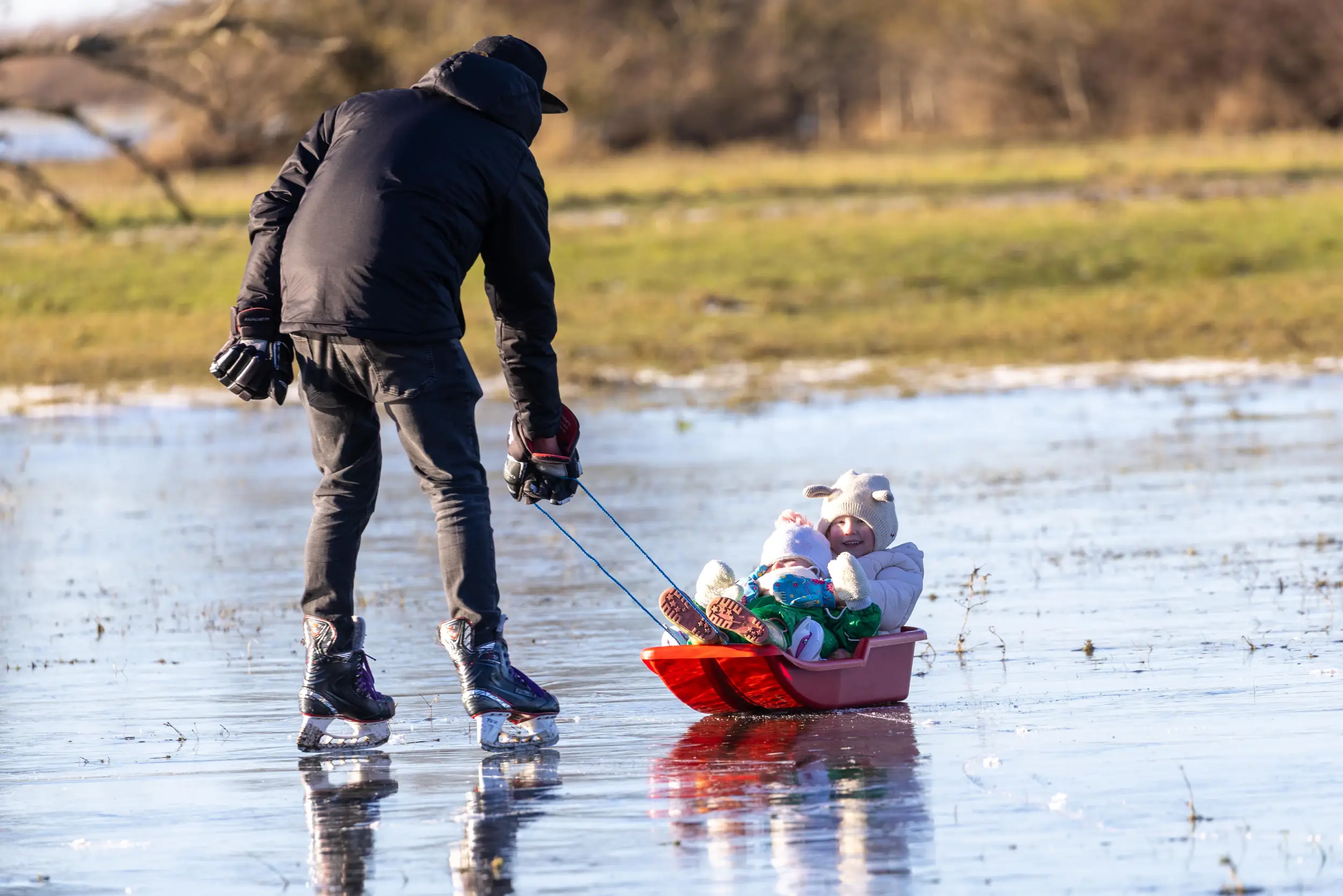 An adult skating on a frozen Fenland waterway while pulling a child on a sledge